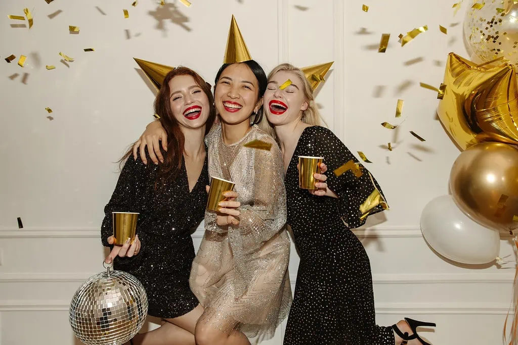 Three friends laughing at a party with hats, golden cups, confetti, and balloons
