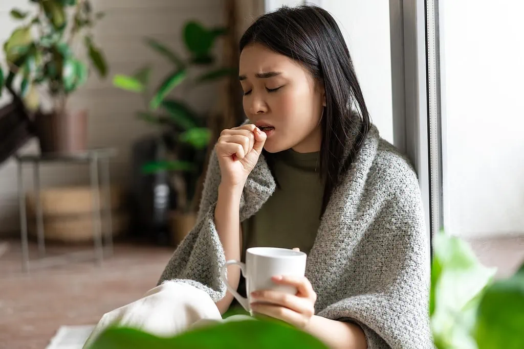Asian woman coughing into hand, wrapped in blanket, holding mug.